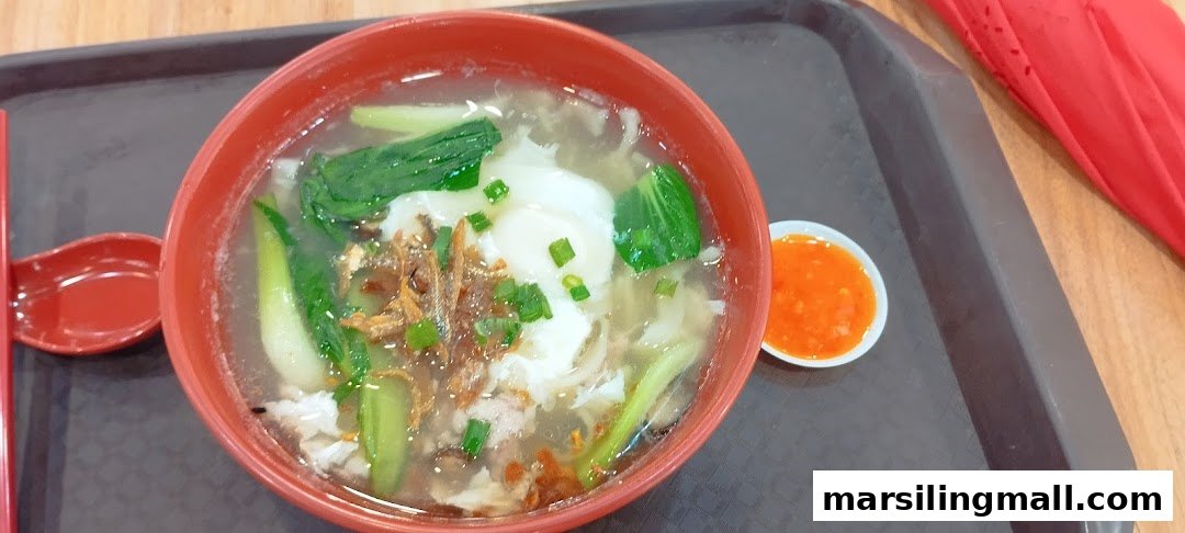A close-up of a hearty bowl of clear noodle soup with vegetables, egg, and fried anchovies, served with chili.