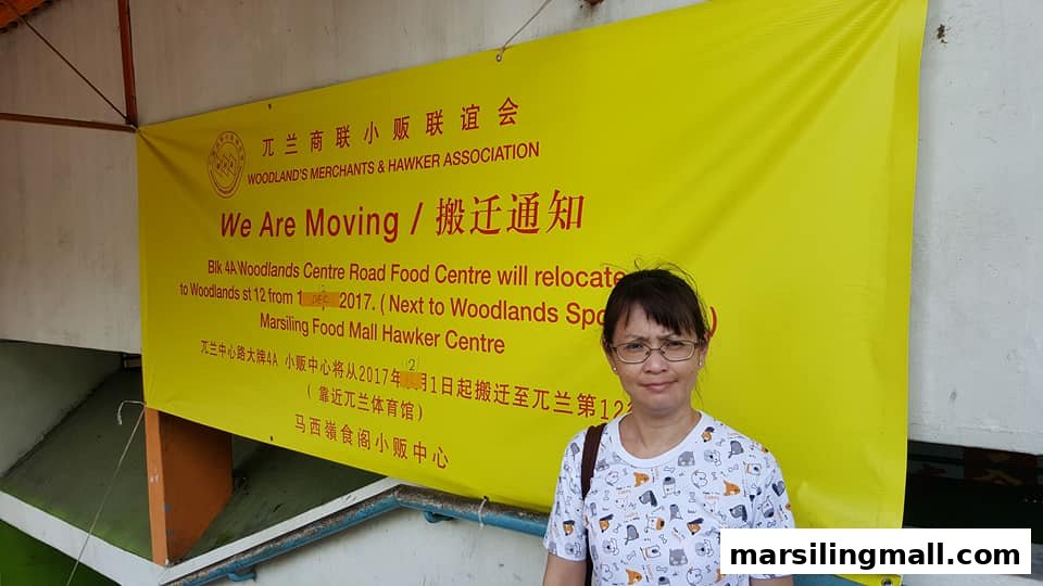 A woman stands before a yellow banner announcing the relocation of Woodlands Centre Road Food Centre.