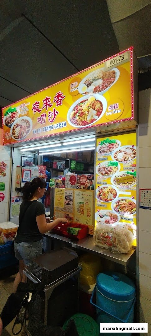 A vibrant food stall, 'Ye Lai Xiang Laksa', with a customer at the counter.