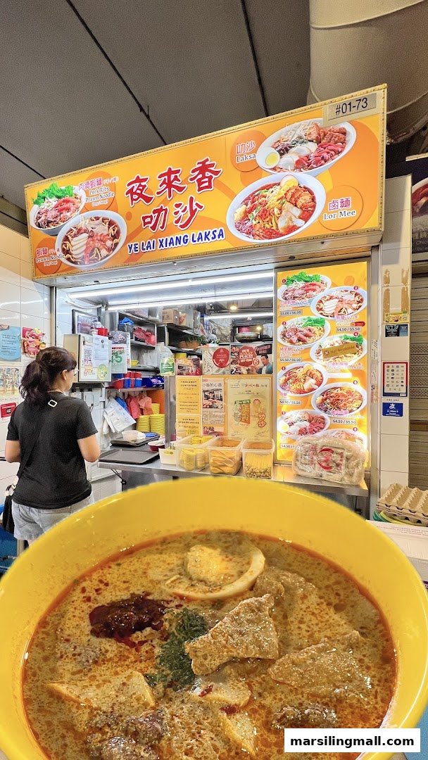 A customer orders at Ye Lai Xiang Laksa stall, with a vibrant bowl of laksa in the foreground.