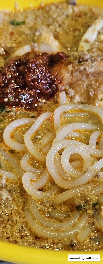 A close-up of rich, flavorful laksa noodles with a dollop of spicy chili paste in a yellow bowl.