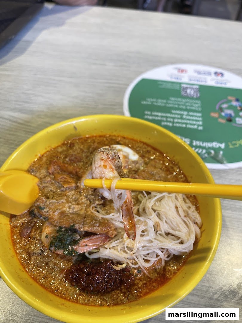 A vibrant bowl of Laksa with prawns and noodles, held by yellow chopsticks, on a table.