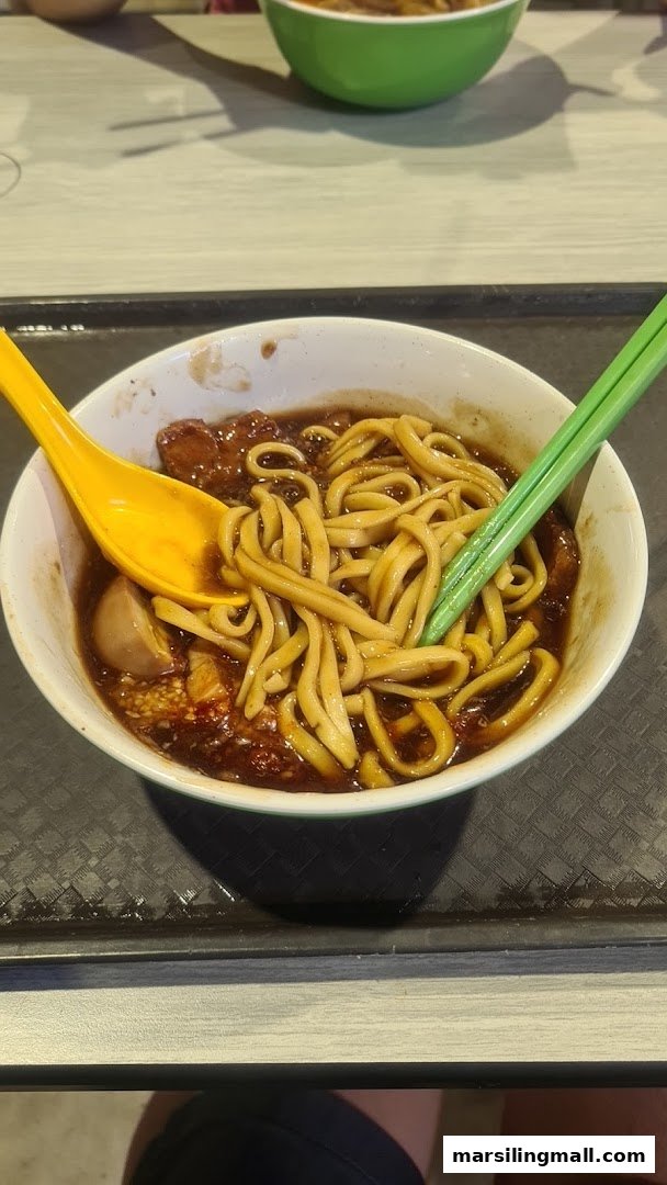A close-up of a delicious bowl of dark sauced noodles with a spoon and chopsticks.