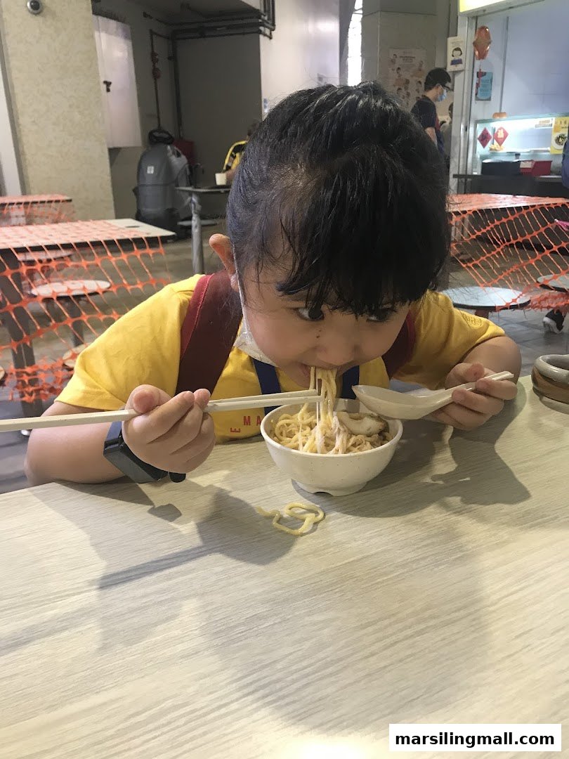 A child in a yellow shirt enthusiastically eats a bowl of noodles with chopsticks and a spoon.