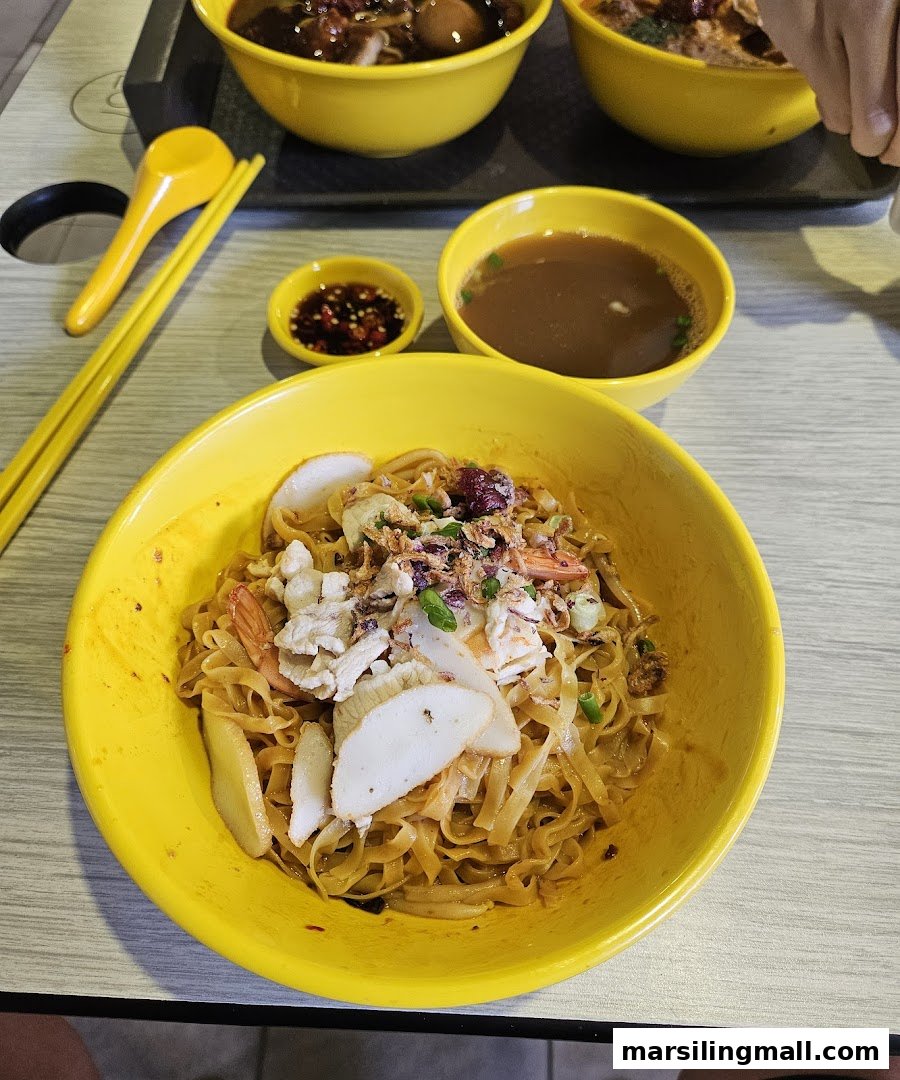 A vibrant yellow bowl of delicious dry noodles with seafood and fried shallots, accompanied by soup and chili.