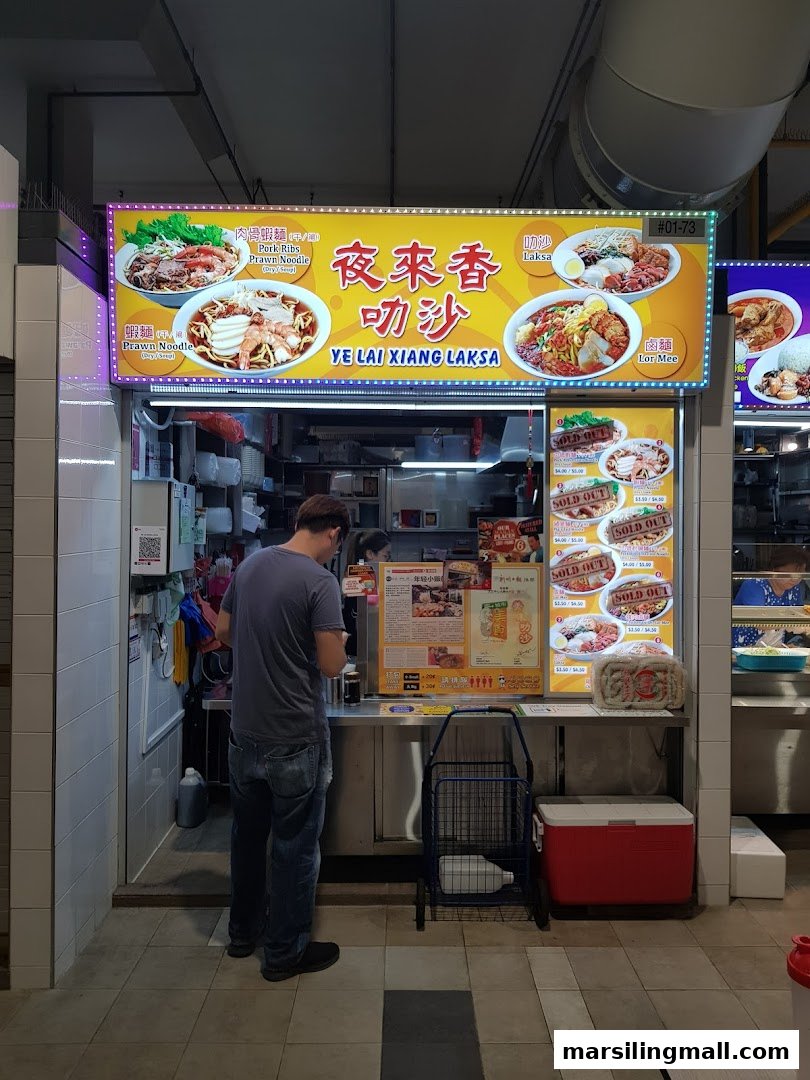 A customer stands at the counter of Ye Lai Xiang Laksa stall, featuring a bright menu board.