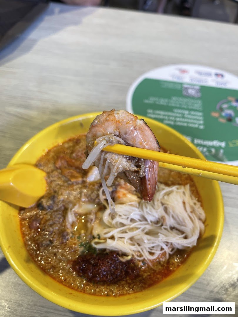 A close-up of a vibrant bowl of laksa with a large shrimp held by chopsticks.