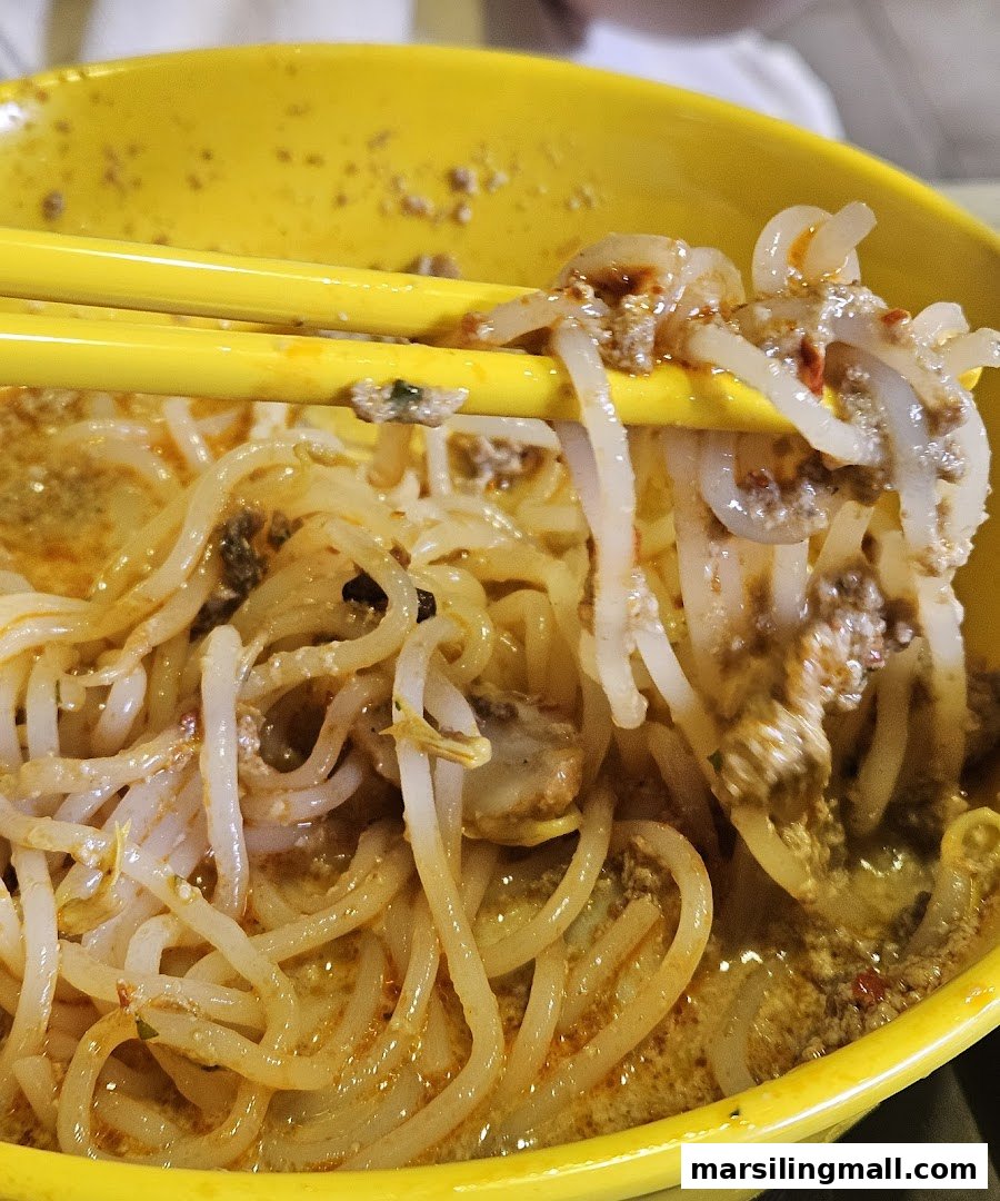 Close-up of laksa noodles and ingredients being lifted from a vibrant yellow bowl with chopsticks.