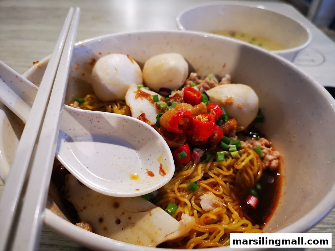 A close-up of a vibrant bowl of minced meat noodles with fish balls, fish cake, and chili.
