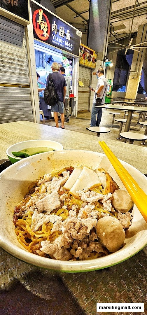 A delicious bowl of minced meat noodles with fish cakes and meatballs, with the Yum-Mee shop in the background.