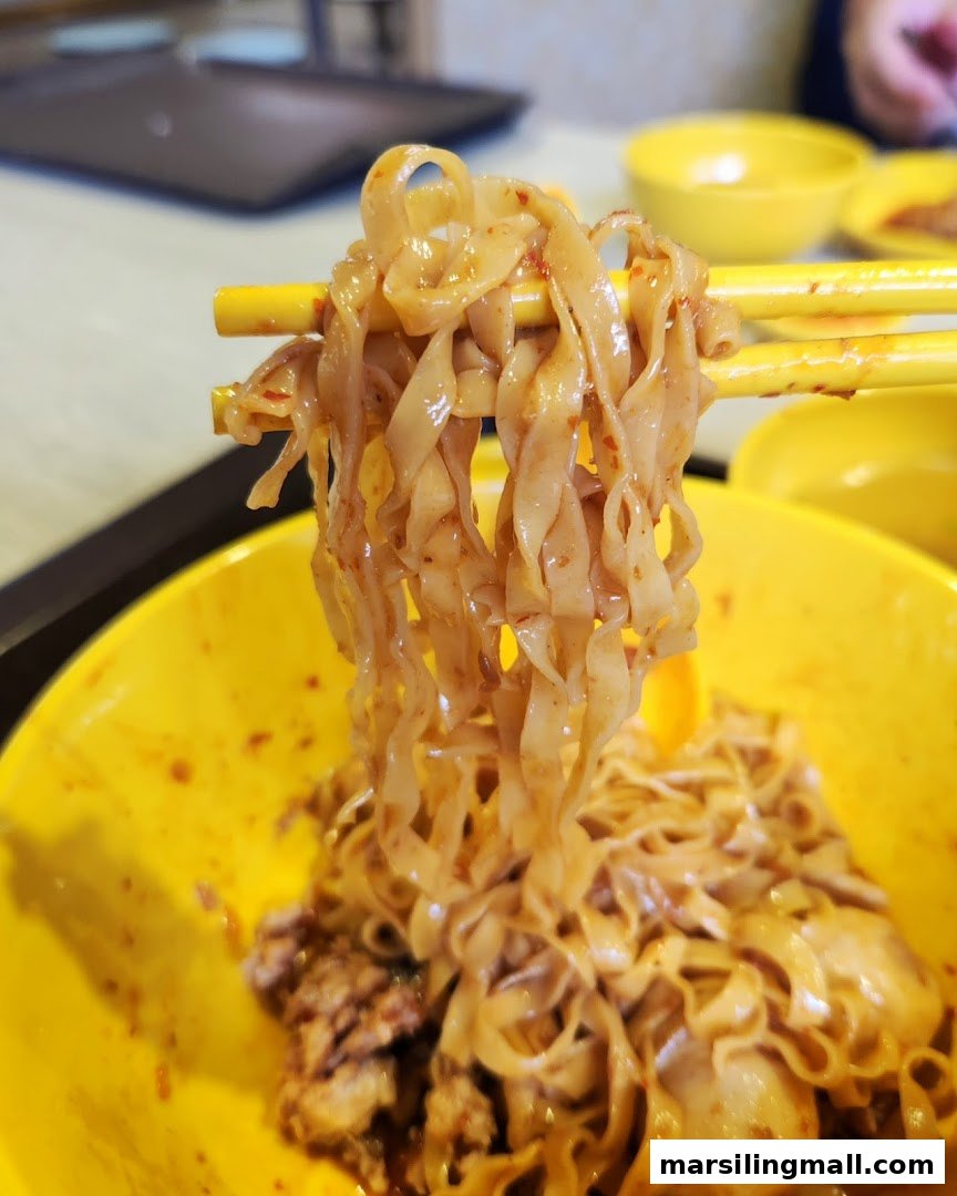 Chopsticks lift saucy flat noodles from a vibrant yellow bowl, revealing minced meat and fish balls.