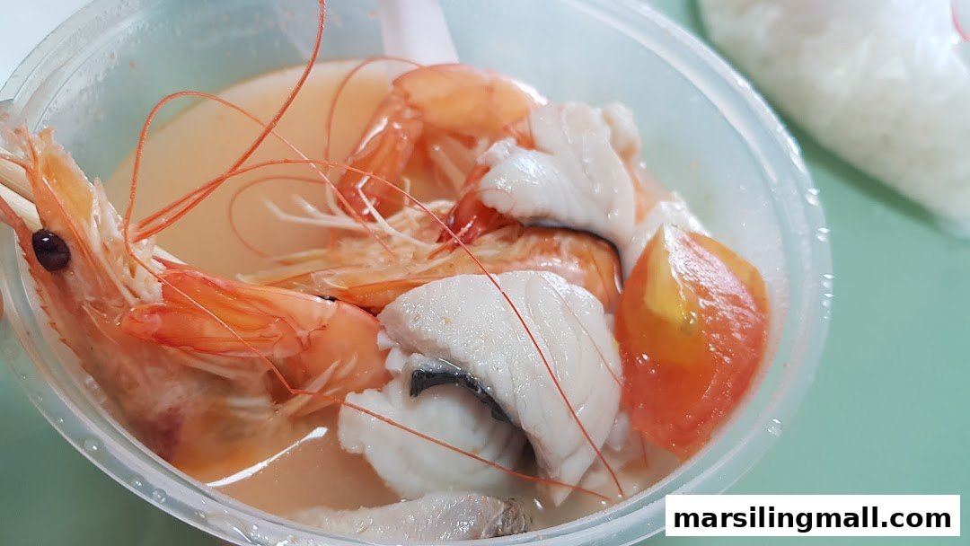 A clear plastic bowl filled with seafood soup, featuring large prawns, fish slices, and tomato.