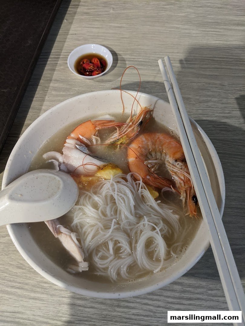 A close-up of a delicious bowl of seafood noodle soup with prawns, squid, and a side of chili sauce.