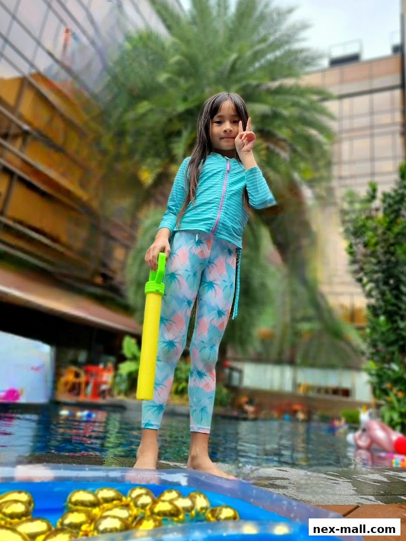 A young girl in swimwear poses by a pool with a water gun and golden balloons.