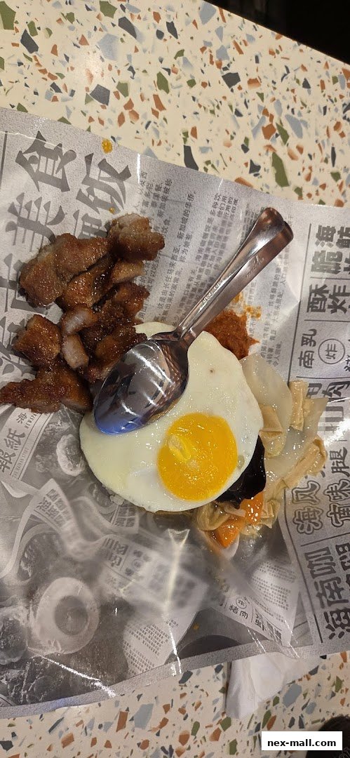 A close-up of a Hainanese dish with fried pork, a sunny-side-up egg, and vegetables on newspaper-style wrapping.
