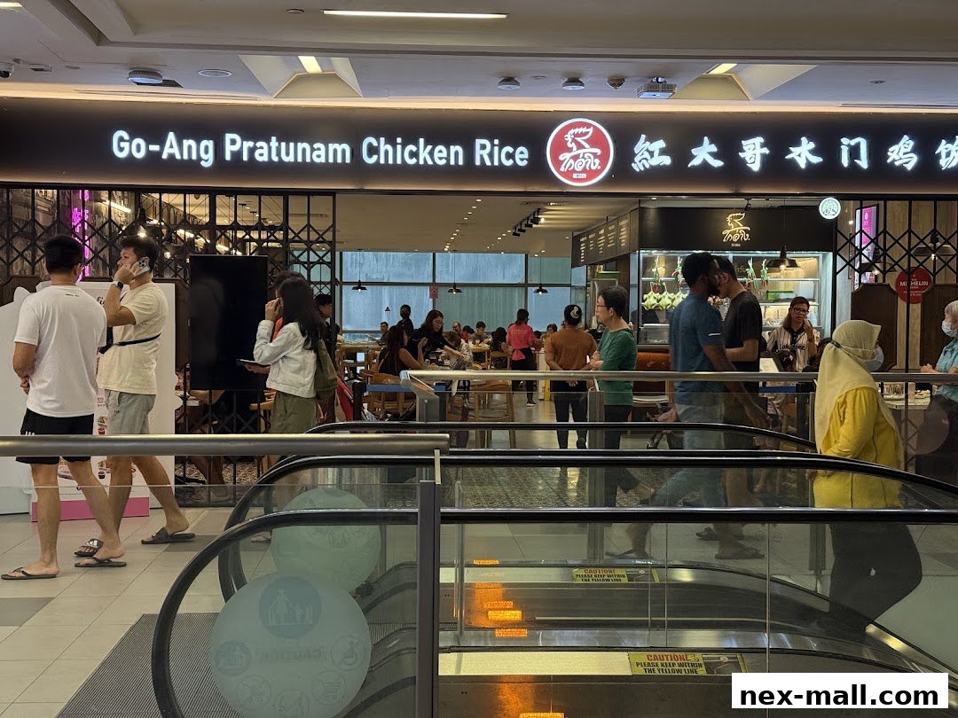 The exterior of Go-Ang Pratunam Chicken Rice restaurant with customers inside and on escalators.