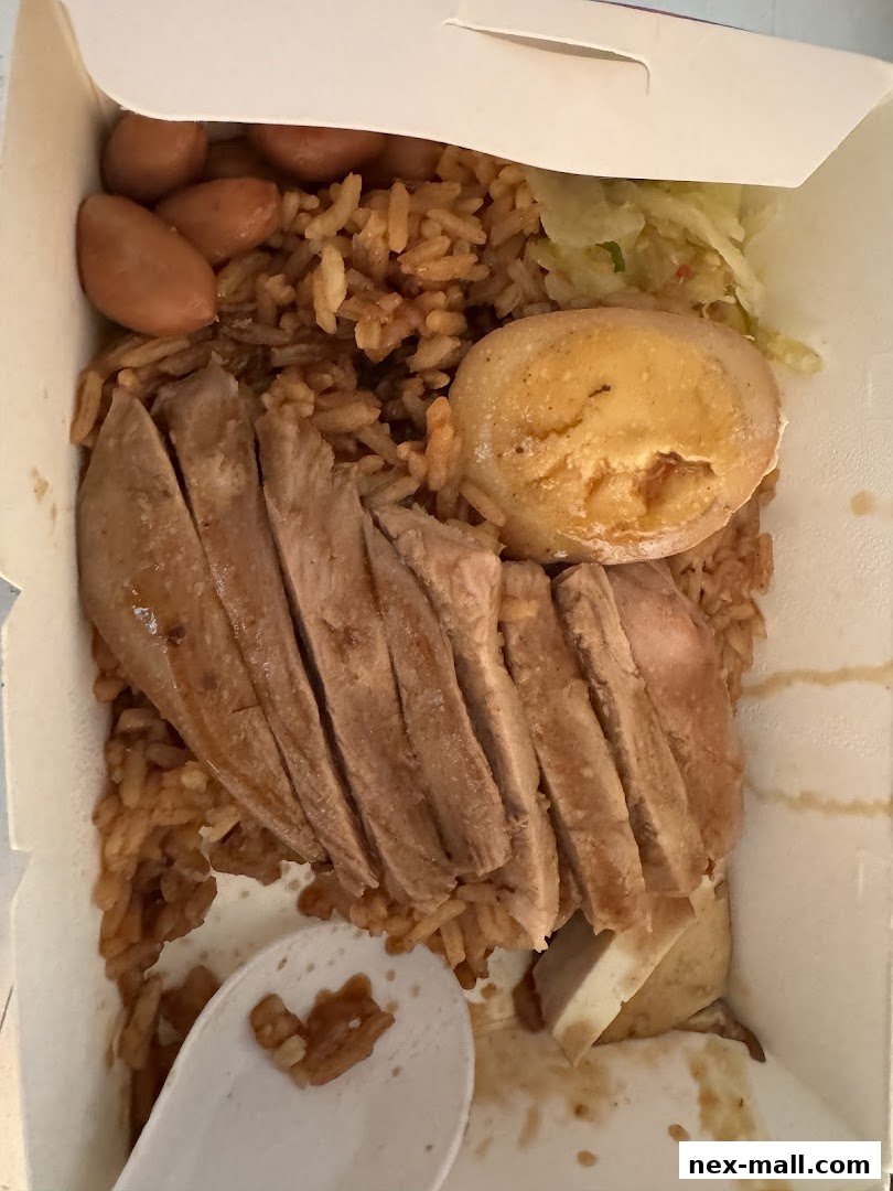 A takeout box filled with rice, sliced meat, sausages, a hard-boiled egg, and pickled vegetables.