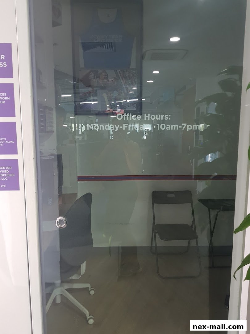 The glass entrance of Anytime Fitness NEX, showing office hours and interior reflections.