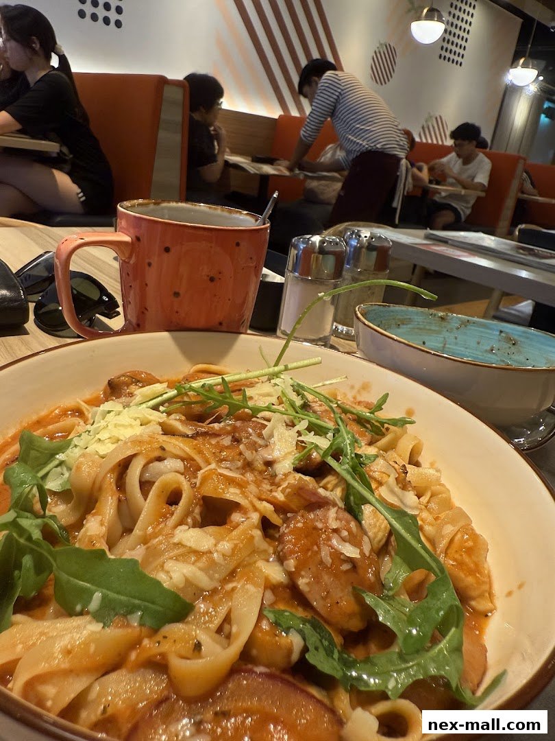 A close-up of a pasta dish with a mug and people dining in the background.