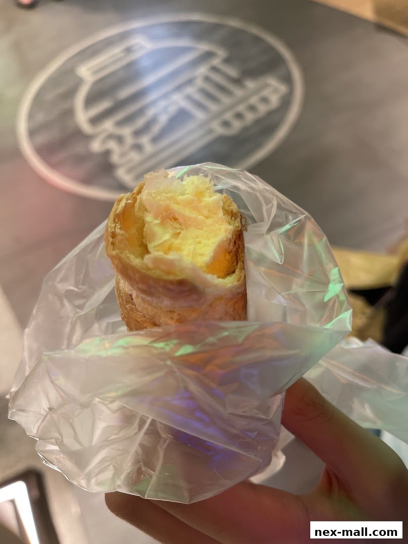 A hand holds a pastry filled with creamy custard, wrapped in plastic.