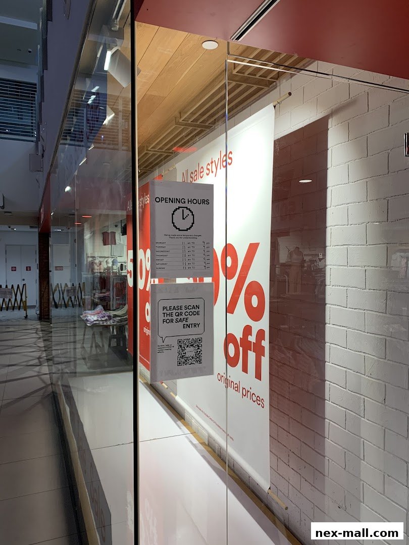 The storefront of Cotton On NEX features large sale banners and an opening hours sign.