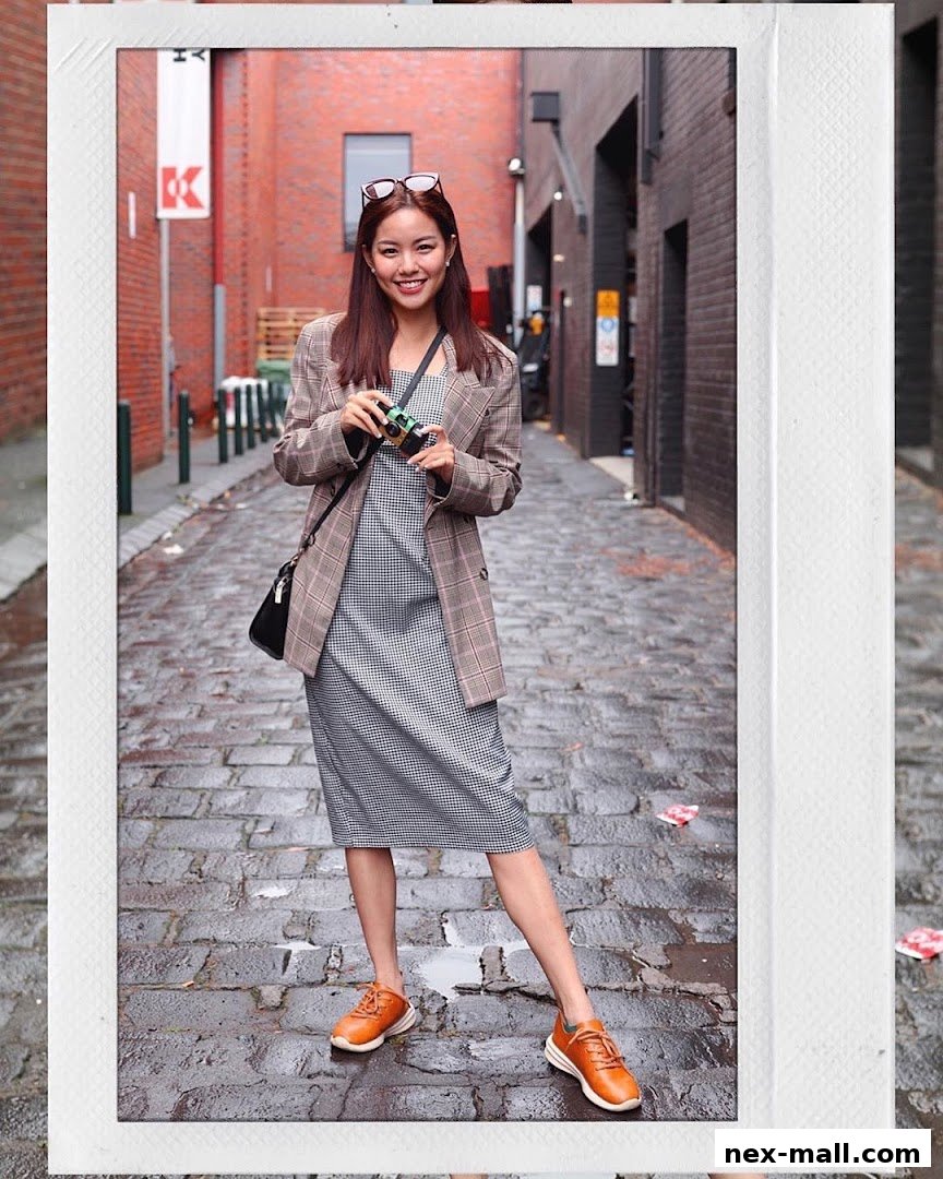 A smiling woman in a checkered dress and blazer holds a camera in a cobblestone alley.