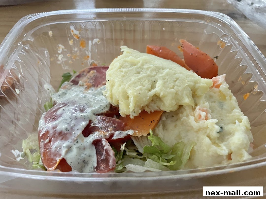 A clear plastic container filled with a salad featuring mashed potatoes, tomatoes, carrots, and lettuce with dressing.