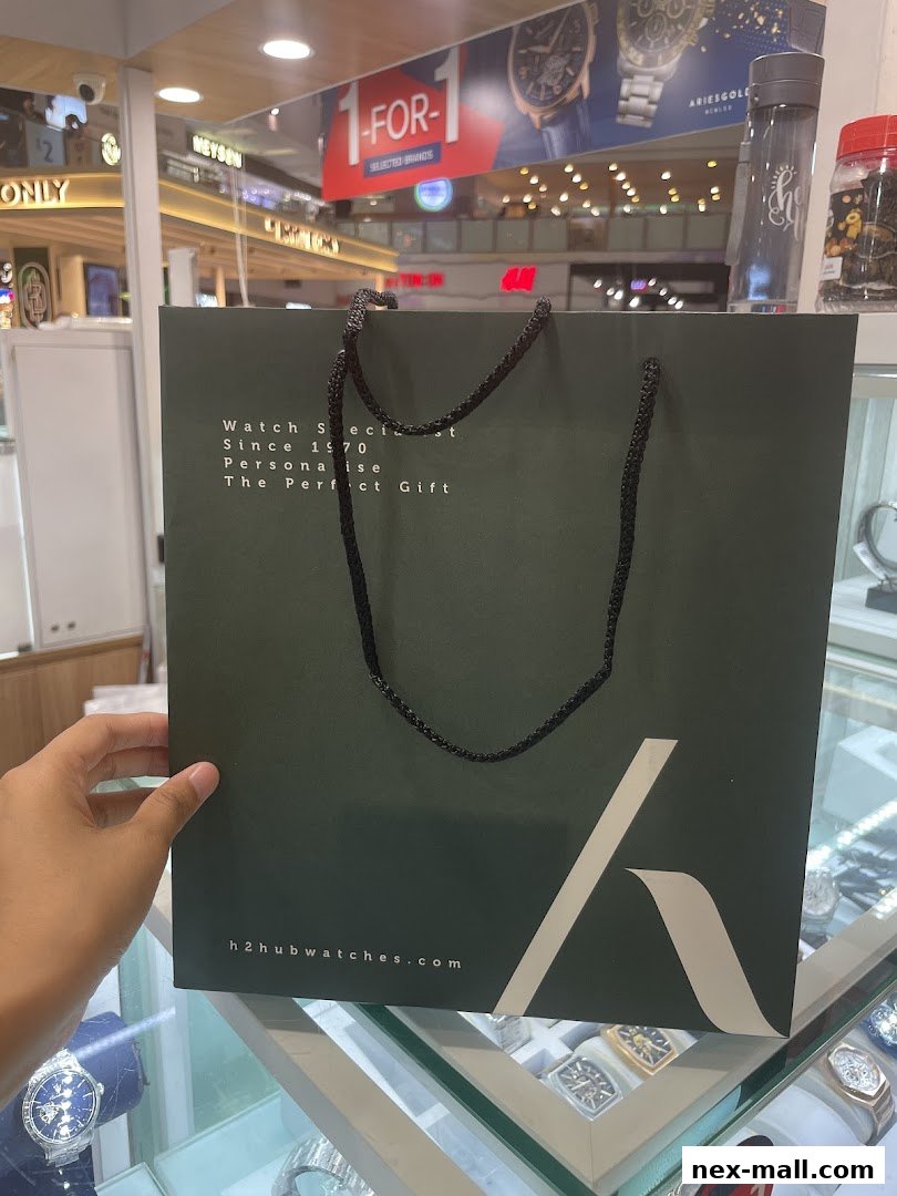 A person holds a dark green shopping bag from H2 Hub Timepiece with watch-related text and logo.