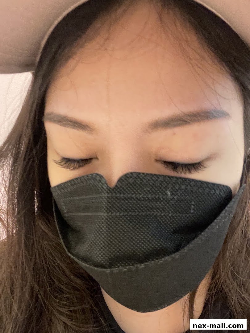 A close-up of a woman with eyelash extensions, wearing a black face mask and a light-colored headband.