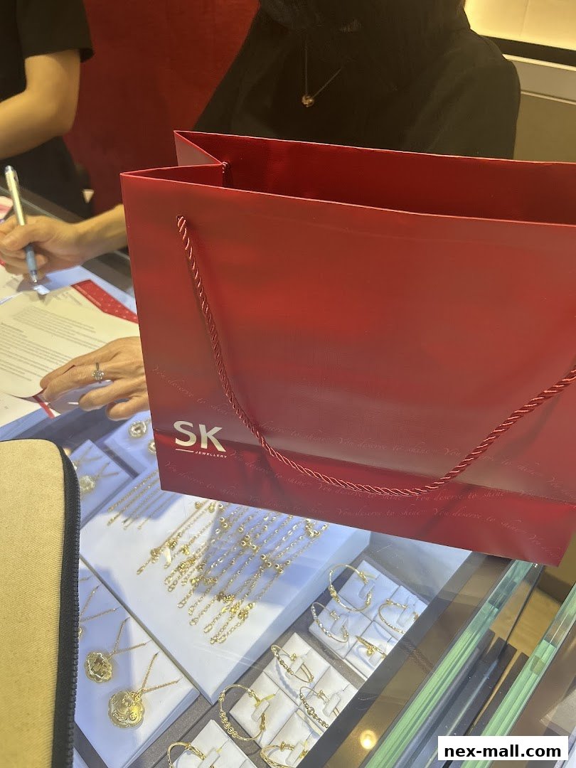 A red shopping bag with SK Jewellery logo sits on a display of gold necklaces and bracelets.