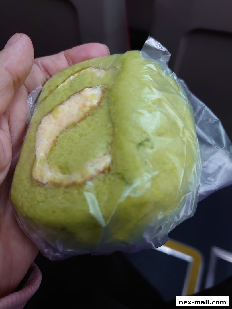 A hand holds a green pandan swiss roll cake wrapped in plastic.