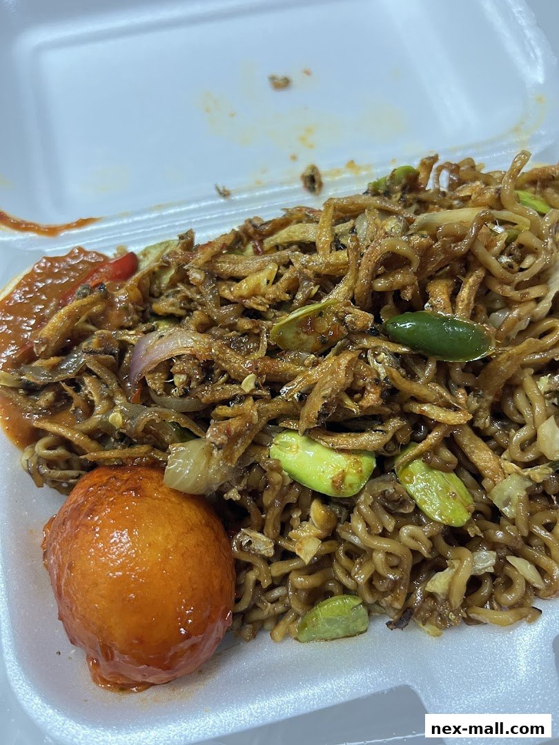 A close-up of a takeout container filled with noodles, anchovies, vegetables, and a fried egg.