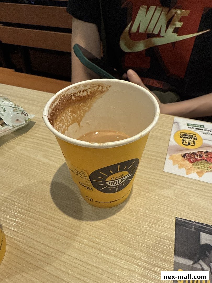 A yellow cup with 'Cafe Hola' branding sits on a wooden table next to a person holding a phone.