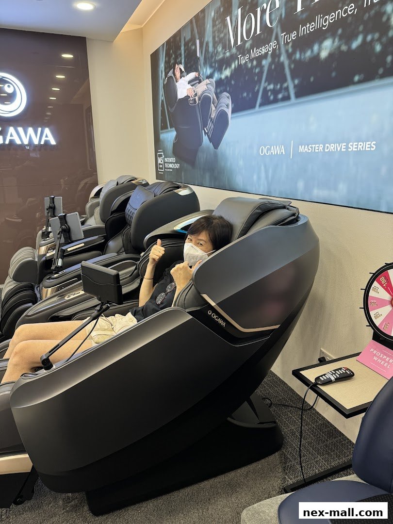 A person sits in an OGAWA massage chair, giving a thumbs up, with other chairs and a promotional wheel visible.