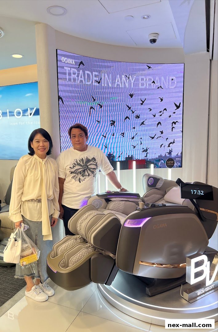 Two people stand next to an OGAWA massage chair with a digital display showing 'TRADE IN ANY BRAND'.