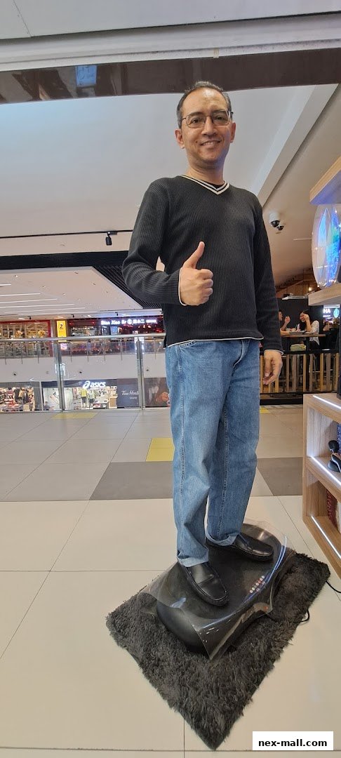 A man stands on a massage device, giving a thumbs-up in a shopping mall.