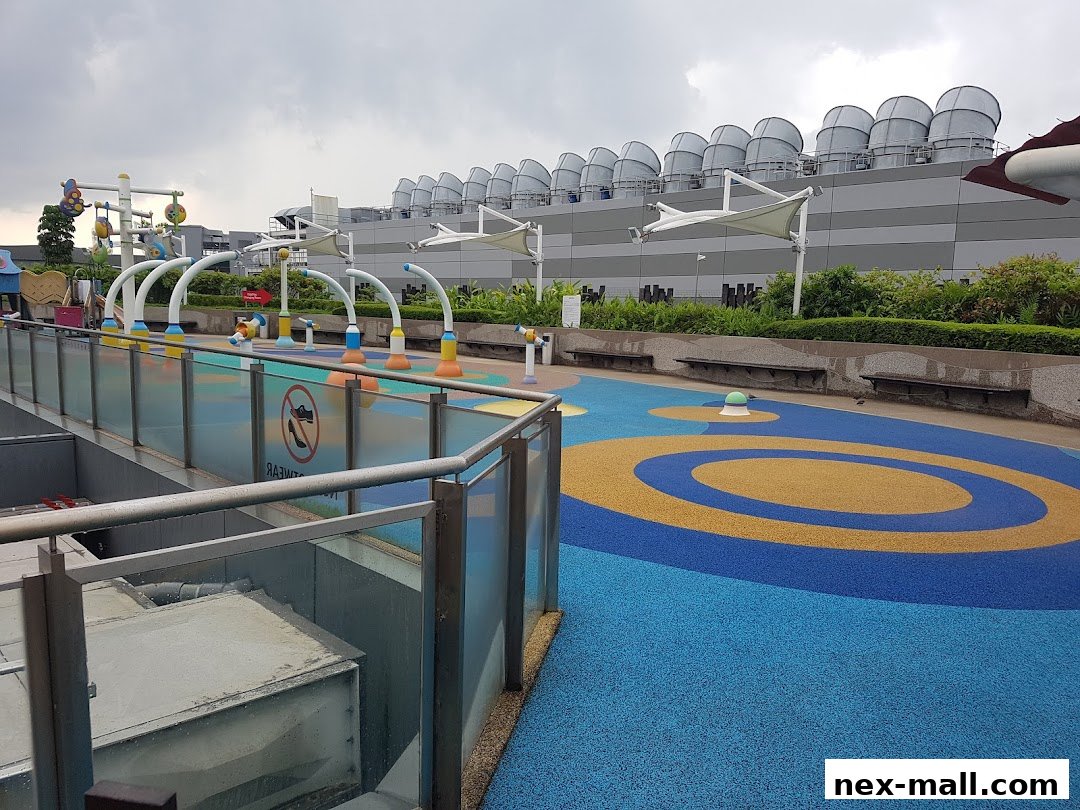 A colorful outdoor water play area with fountains and seating, set against a building with large ventilation units.