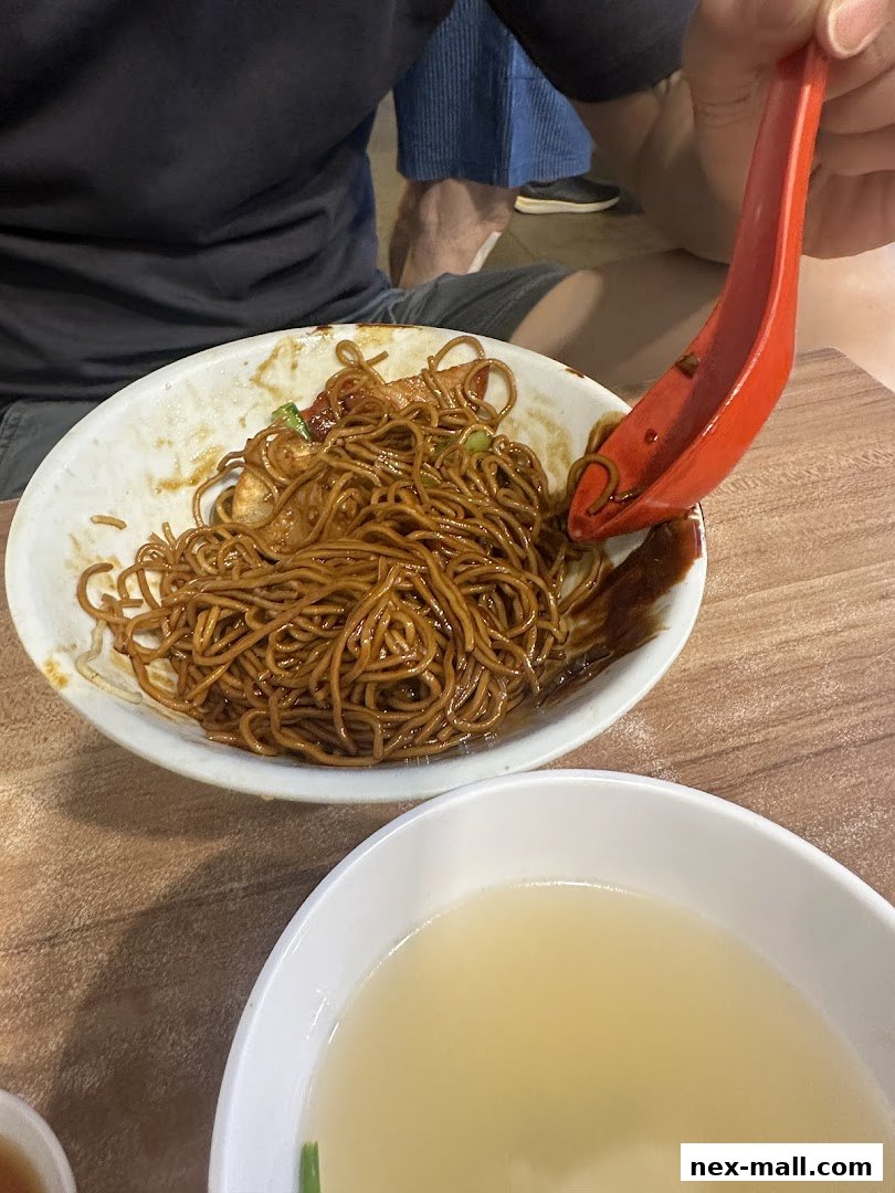 A person is eating a bowl of noodles with a red spoon, with a bowl of soup nearby.