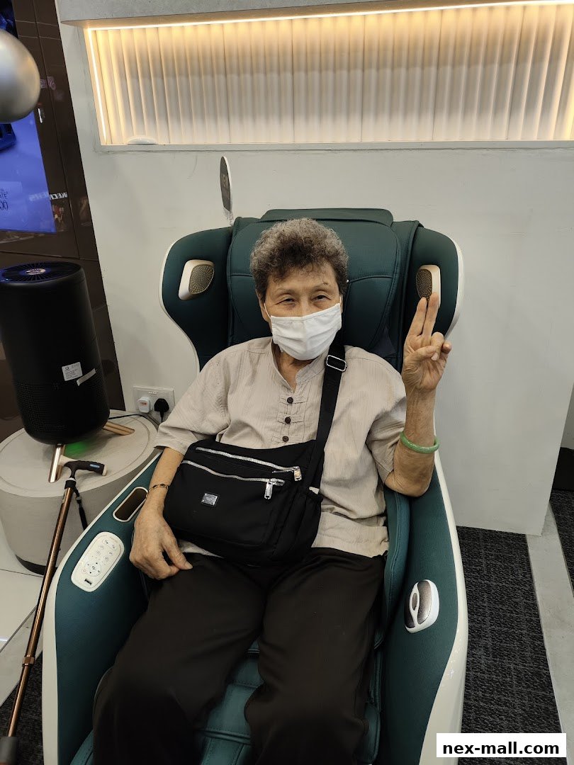 An elderly person wearing a mask sits in a massage chair, giving a peace sign.