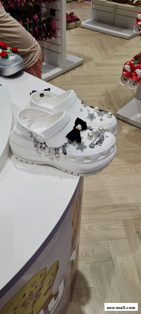 White Crocs decorated with charms, pearls, and a black bow are displayed.