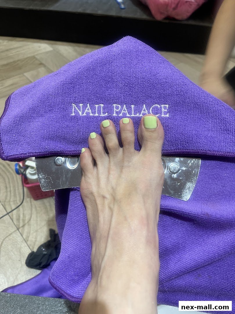A foot with light green nail polish resting on a purple towel with 'Nail Palace' embroidered on it.