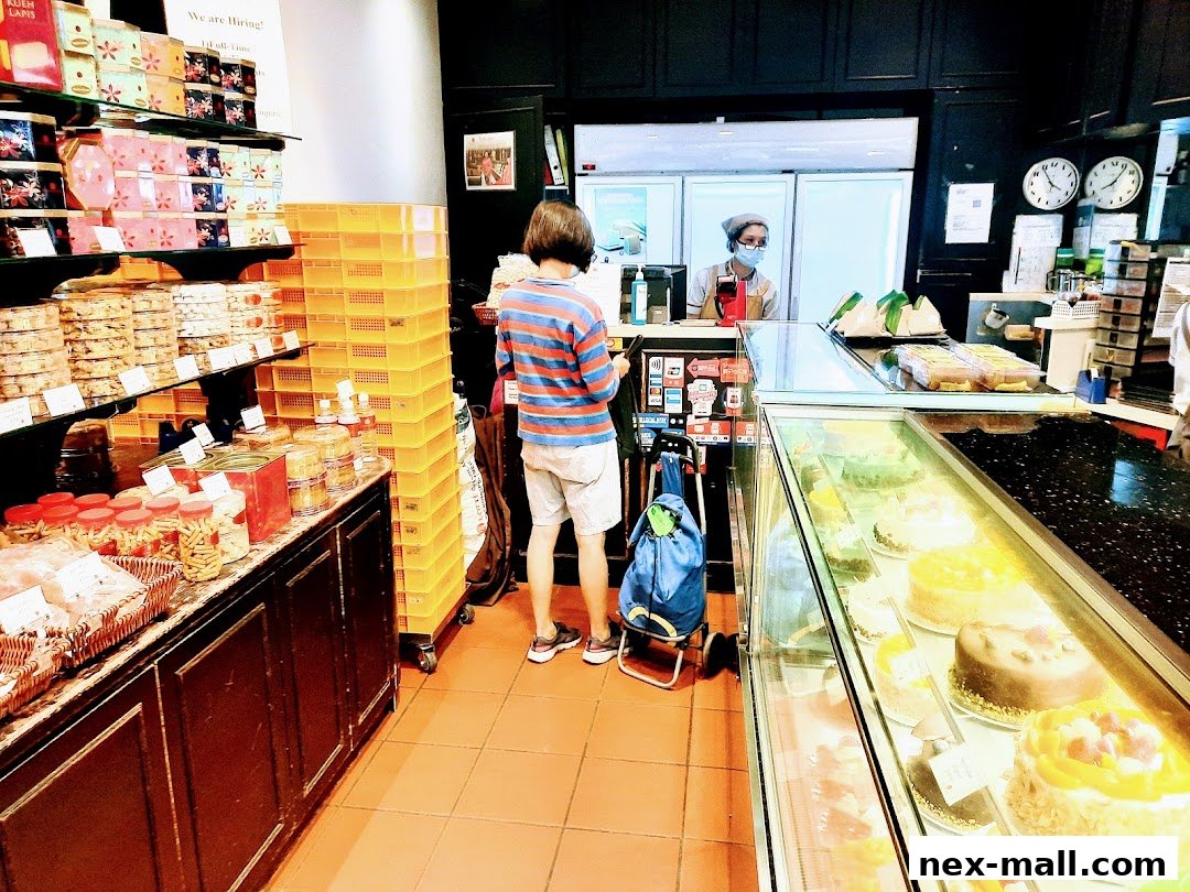 A customer stands in a bakery, browsing cakes and pastries displayed in a refrigerated case.