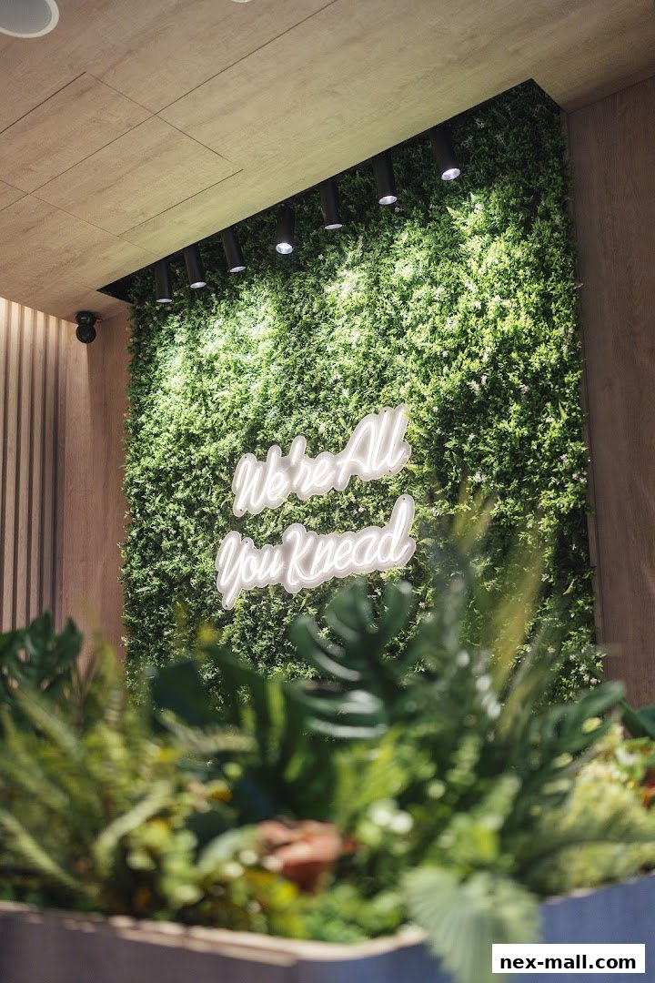 A neon sign saying 'We're All You Knead' on a lush green wall.