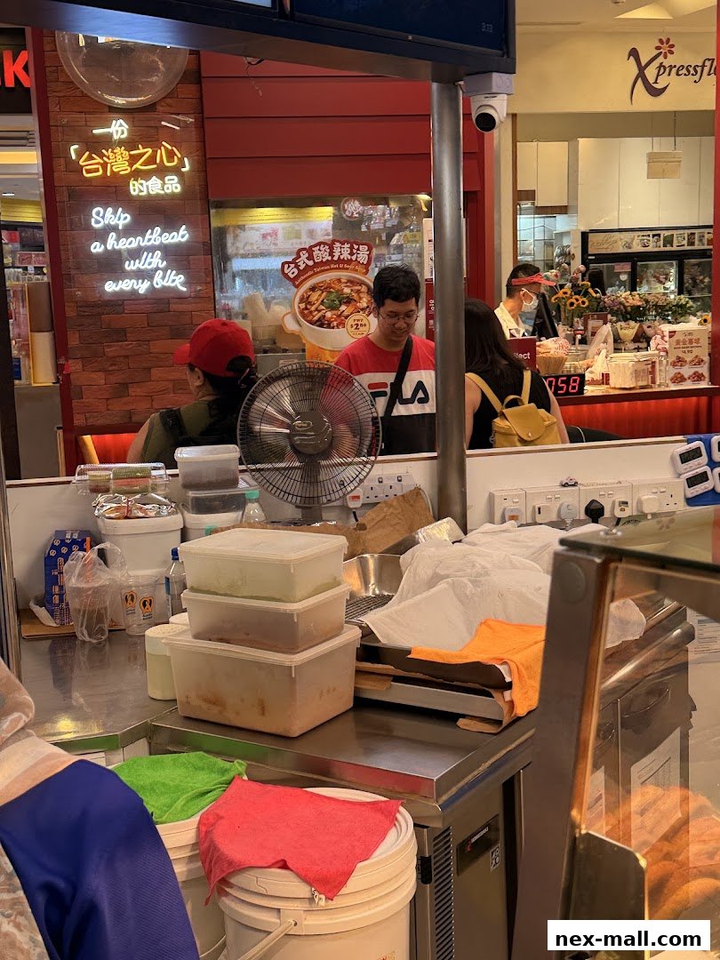A food stall with a neon sign and a display of food items, with customers and staff present.