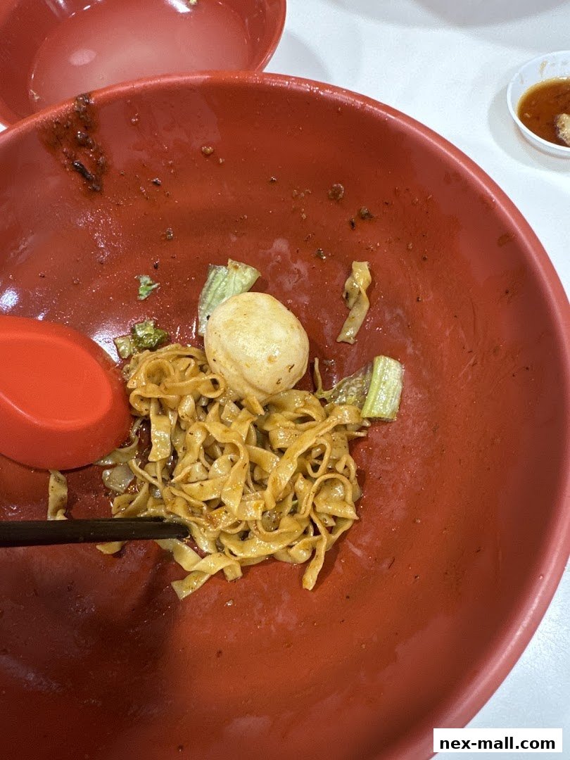 A close-up of a nearly finished bowl of noodles with a meatball and vegetables.