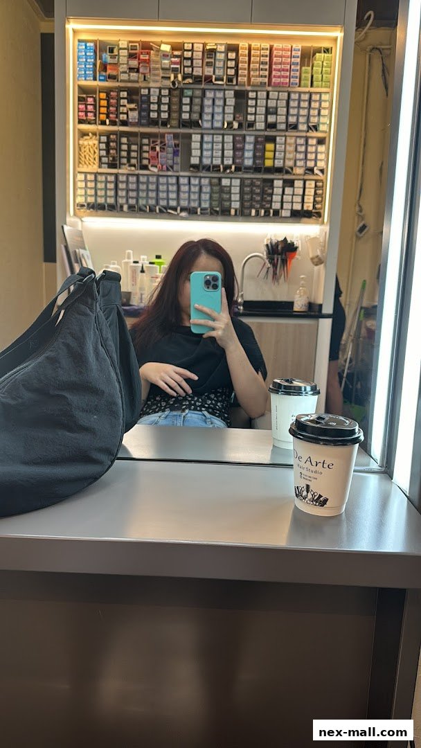 A person takes a selfie in a hair studio, with shelves of hair color products in the background.