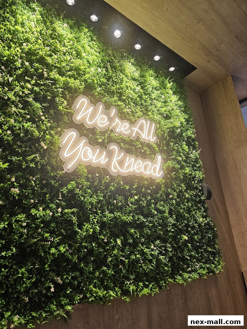 A neon sign saying 'We're All You Knead' on a lush green wall.