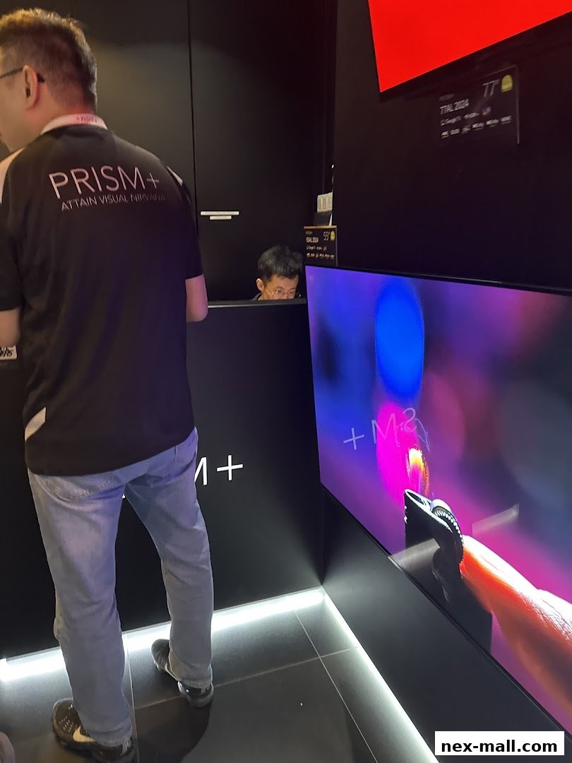 A person wearing a PRISM+ shirt stands near a display with a curved screen showing colorful graphics.
