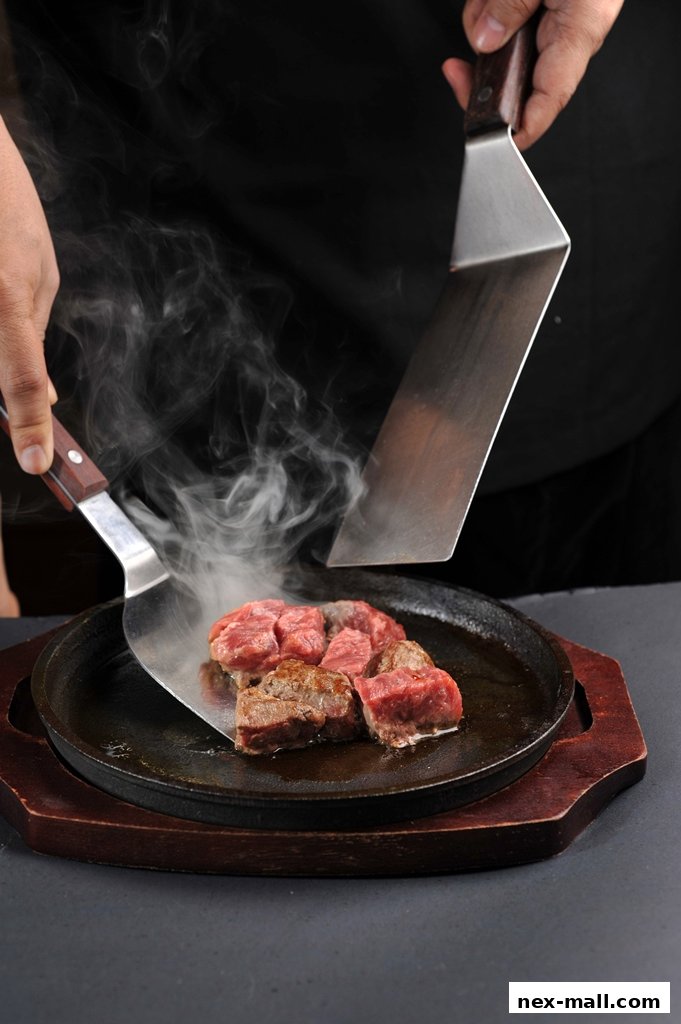A chef is cooking sizzling beef cubes on a hot plate with spatulas.