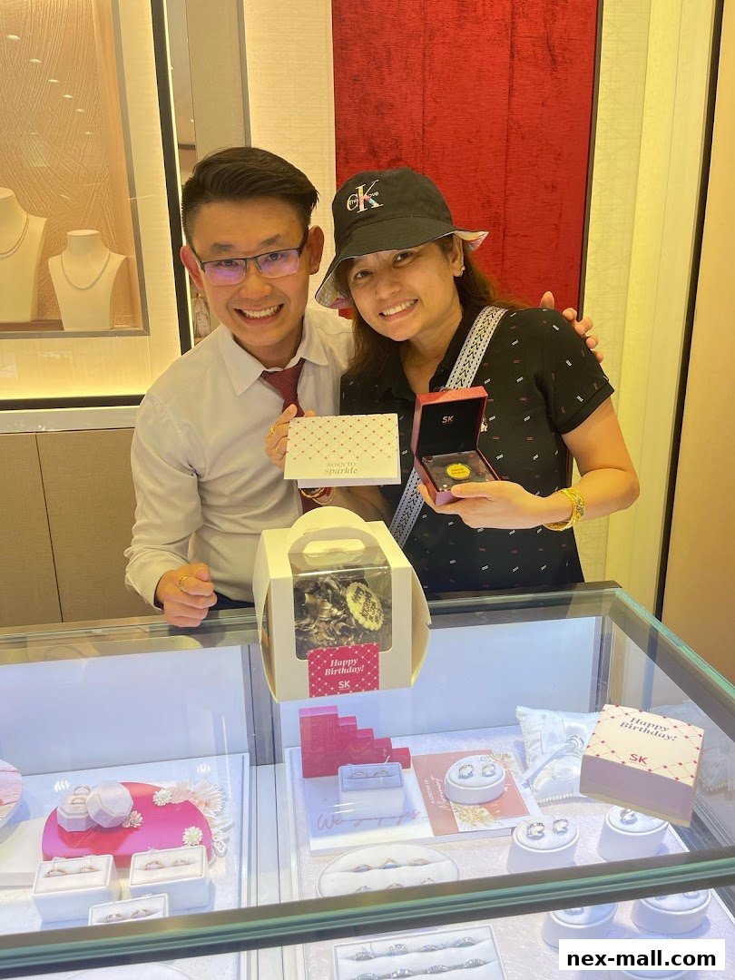 A smiling couple holds jewelry and a birthday cake in a well-lit jewelry store.